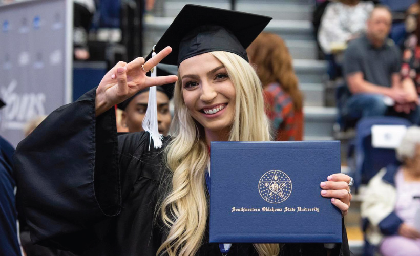 girl with blonde hair wearing graduation cap and gown holding diploma smiling and waiving a peace sign